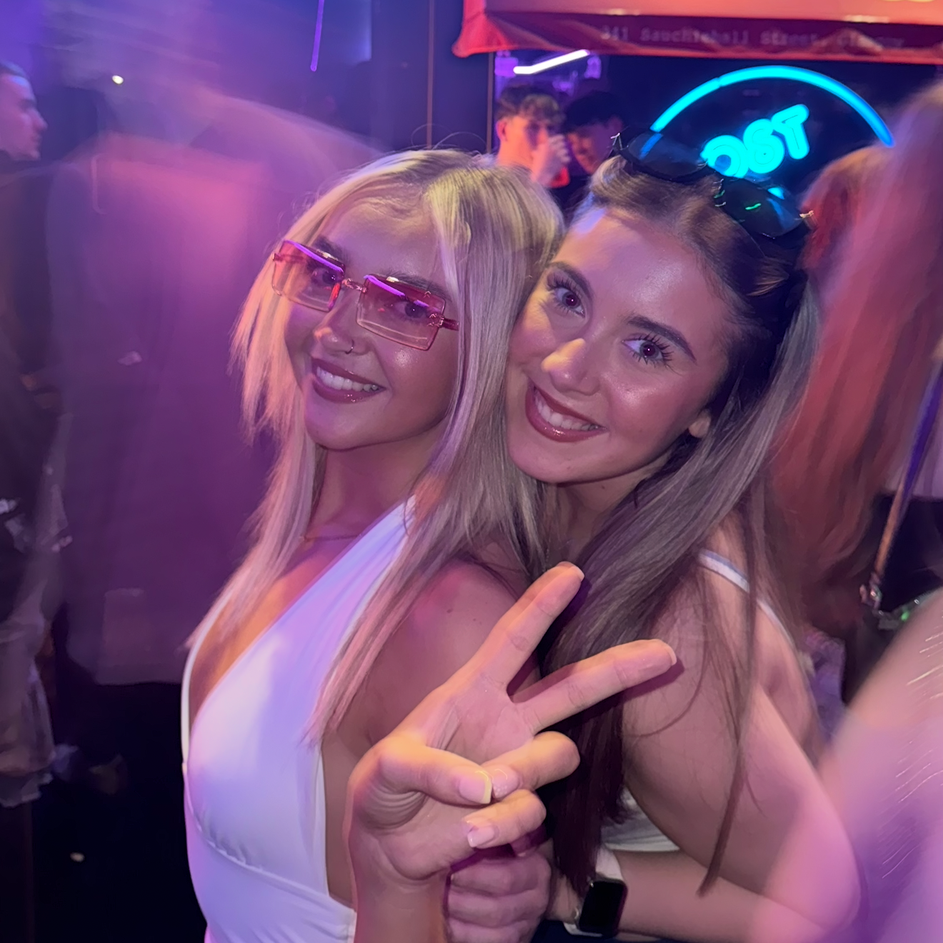 Two women posing together in a nightclub setting with neon signs in the background.