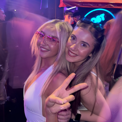 Two women posing together in a nightclub setting with neon signs in the background.